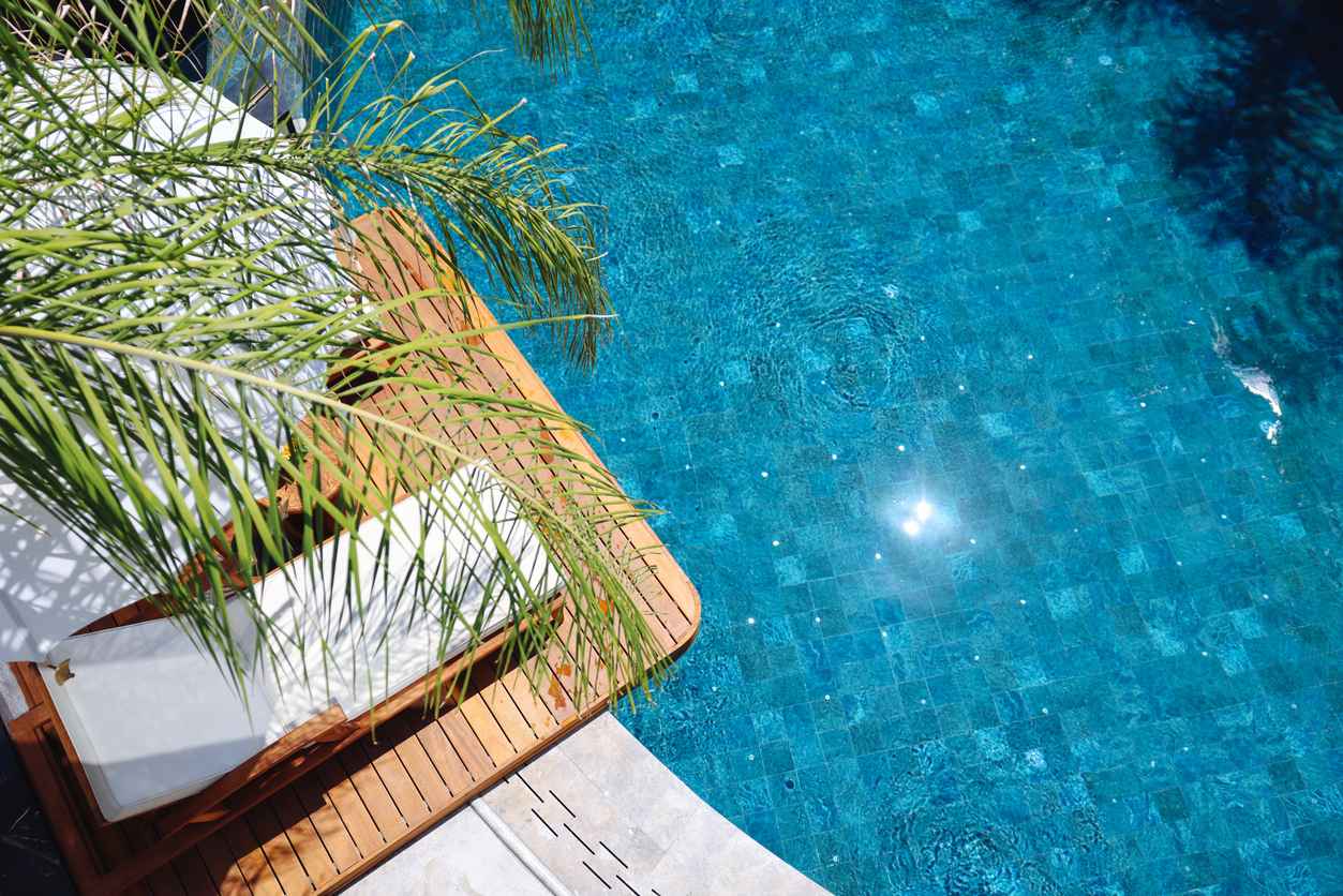 Overview image of two lounge chairs overlooking a backyard pool on a hot sunny day with tropical trees overhanging; Benefits of Backyard Pools