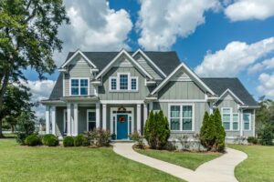 A large gray craftsman new construction House with hardy board siding on a large lot with a green lawn on a clear day with a blue sky and lots of clouds. This home has a lot of curb appeal and exterior shrubs, bushes and flowers.